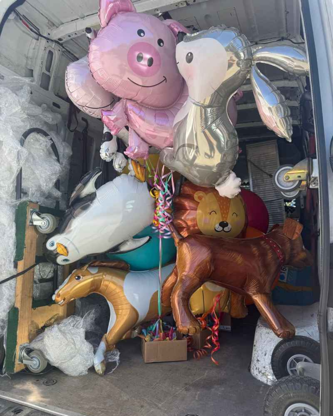 A variety of large animal-shaped foil balloons, including a pig, lion, and horse, inside a Lugg van during a party setup delivery.