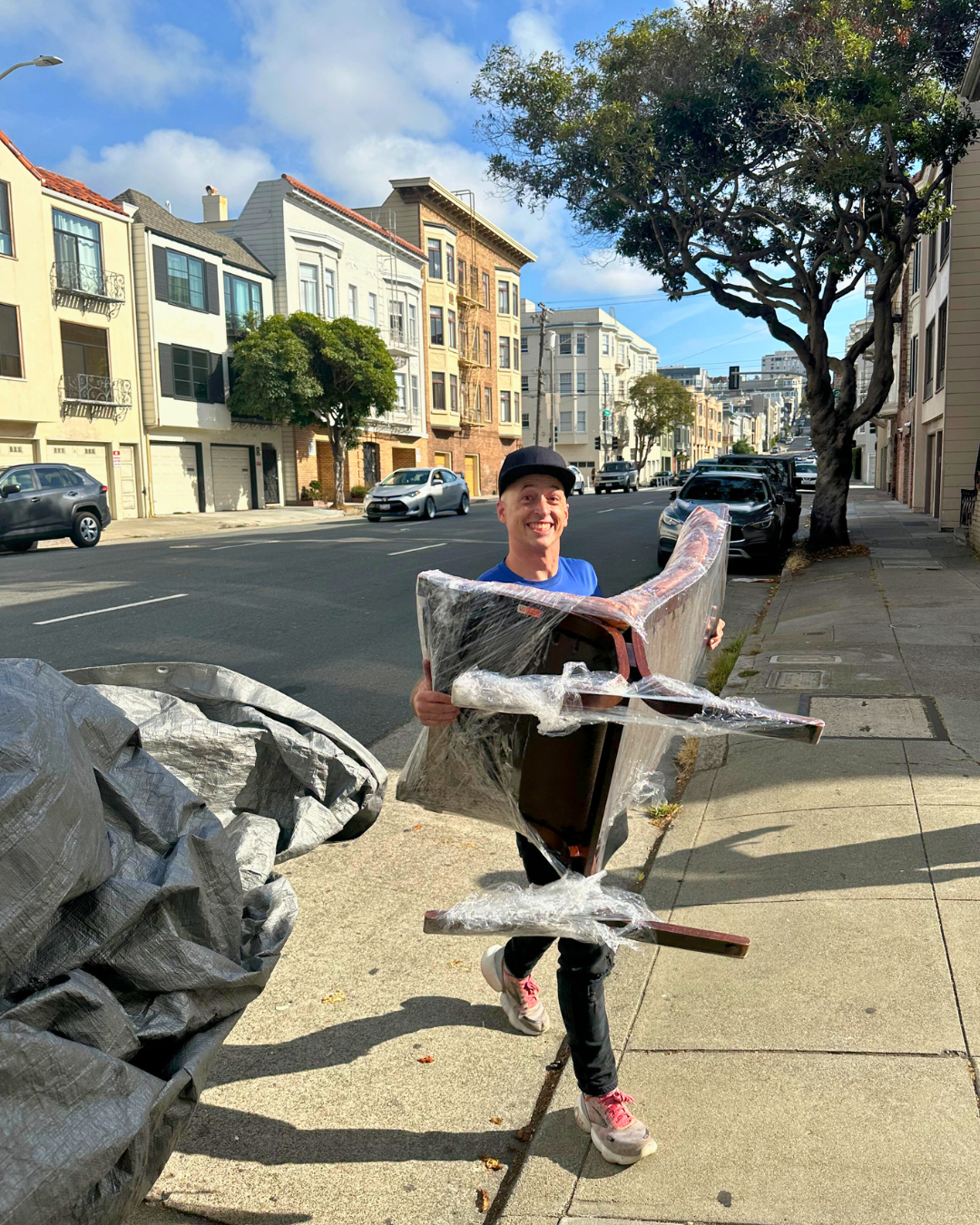 A smiling Lugg mover in a blue shirt and black cap carries a plastic-wrapped mid-century dining chair down a quiet residential sidewalk lined with parked cars and townhouses.