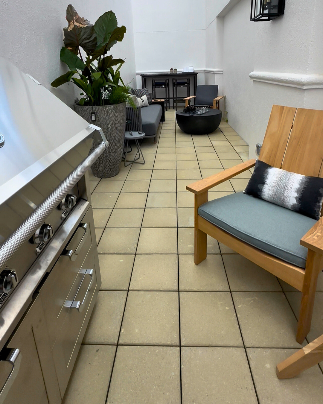 Luxury outdoor patio vignette at Perigold Houston featuring a stainless steel grill, firepit, modern lounge chairs, and potted plants in a narrow courtyard.