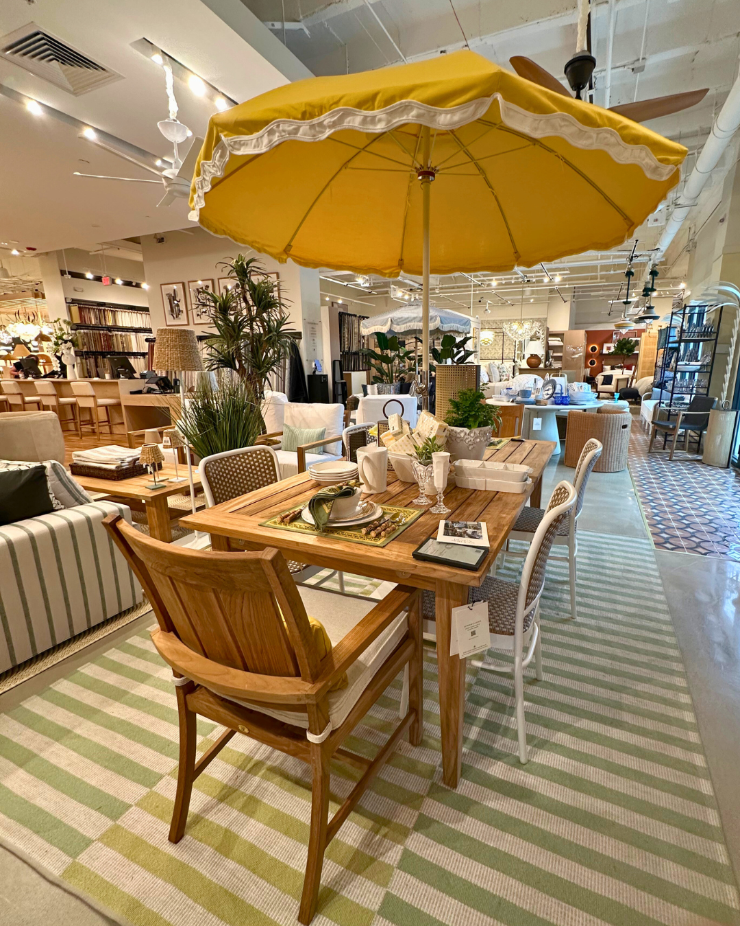 Luxury patio display inside Perigold Houston featuring a yellow umbrella, teak dining table, striped rug, and summer-inspired place settings.