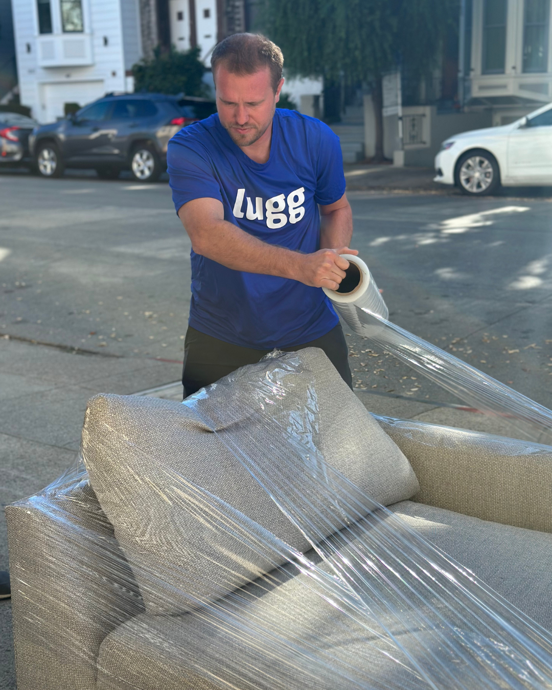 Lugg mover wrapping a light-colored couch with stretch film outside on the street.