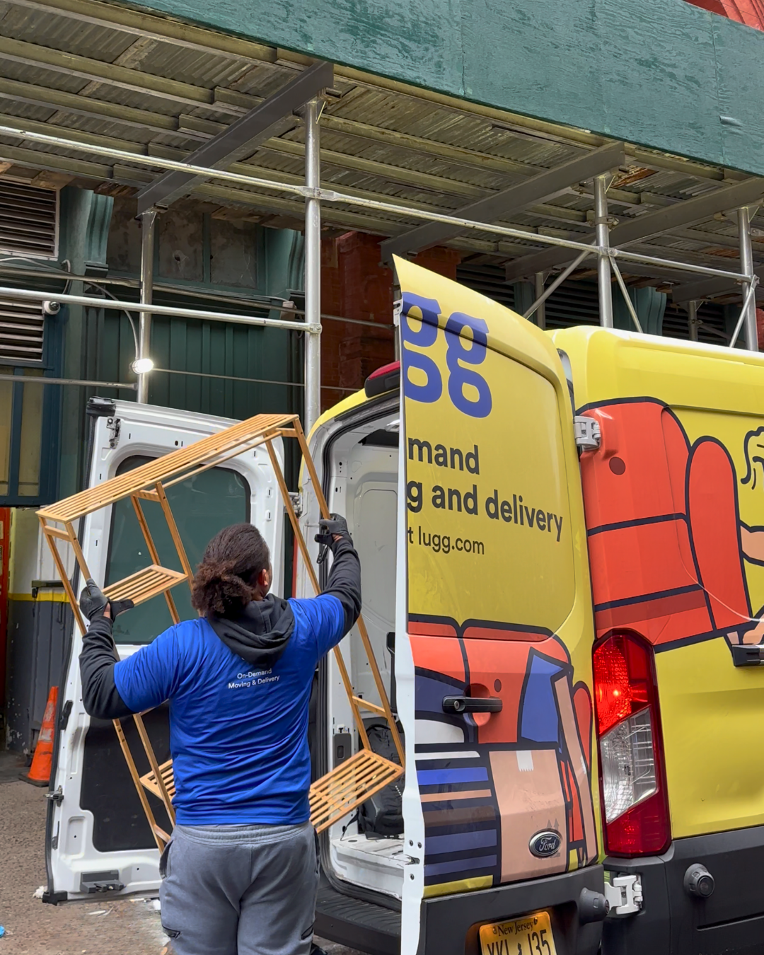 A mover in a blue Lugg shirt lifting a wooden shelf from the back of a branded yellow van on a busy Brooklyn street