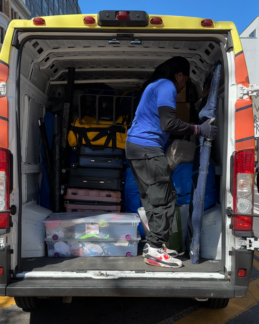 A Lugg mover in blue shirt organizing furniture and boxes inside a packed van on a sunny NYC day.