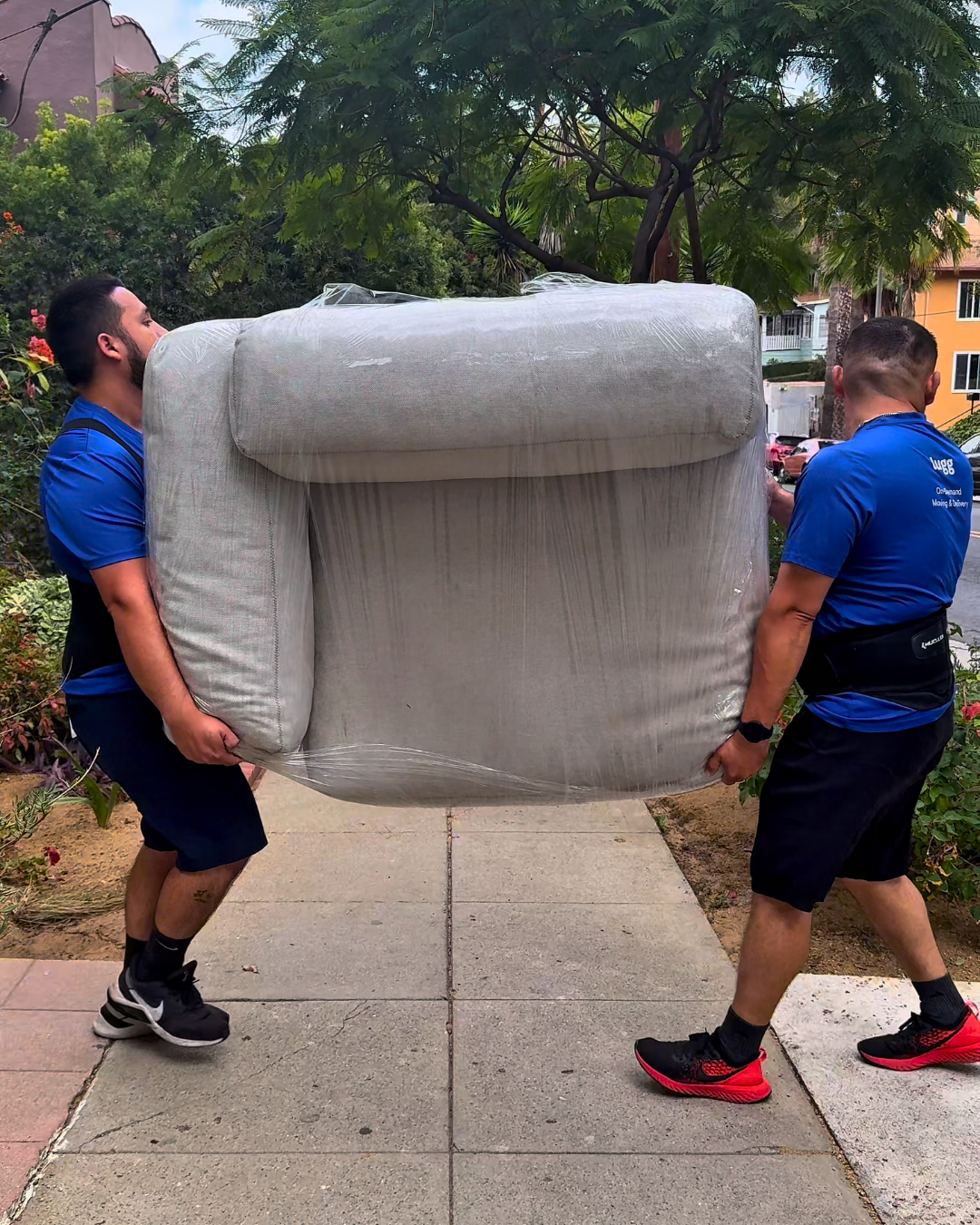 Two Lugg movers in blue shirts carry a wrapped sofa outside, showing how teamwork and small early steps can make moving day smoother.