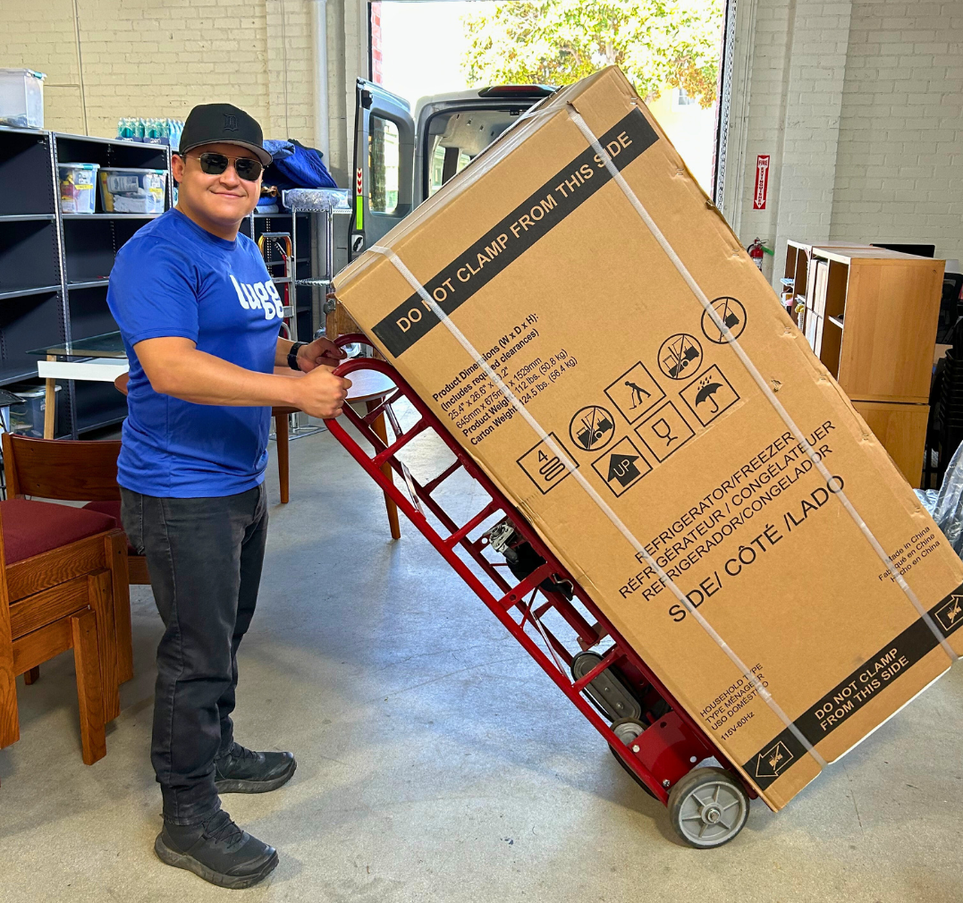 A Lugger in a Lugg shirt transports a large boxed refrigerator using a red appliance dolly inside a warehouse.