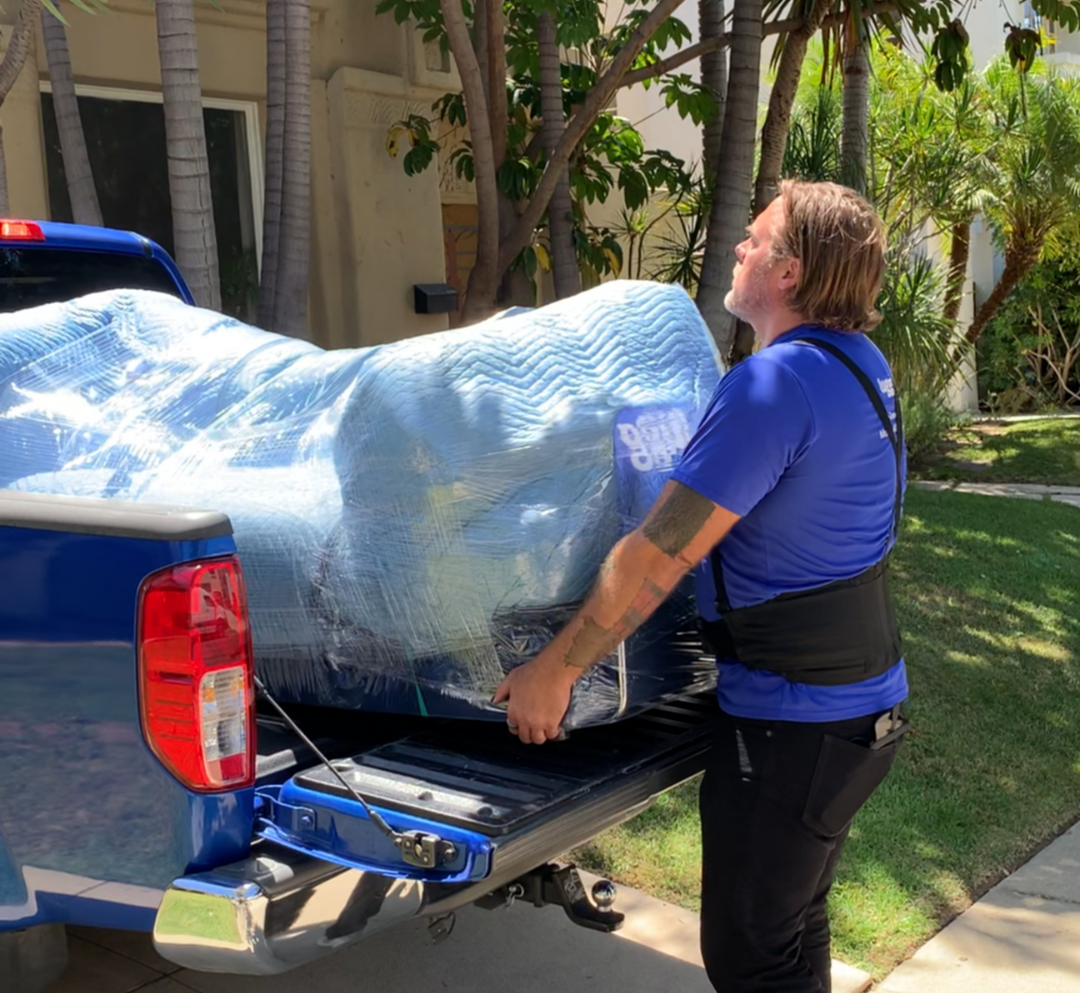 Lugg mover in blue shirt loading a fully wrapped couch into the bed of a blue pickup truck in a residential driveway.
