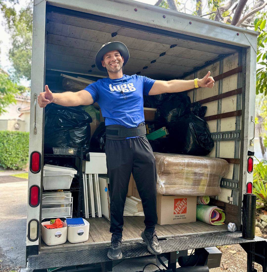Lugg mover in blue shirt and hat standing with arms outstretched in the back of a fully loaded moving truck.