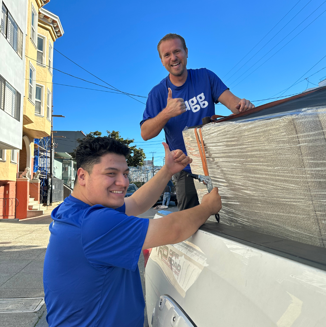 Two Lugg movers smiling and giving thumbs up while securing a wrapped couch in the back of a pickup truck.