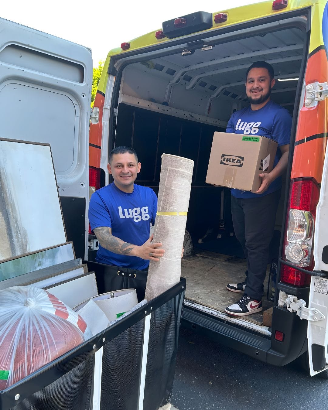Two smiling Lugg movers in blue shirts holding boxes and a rolled rug at the back of a moving van
