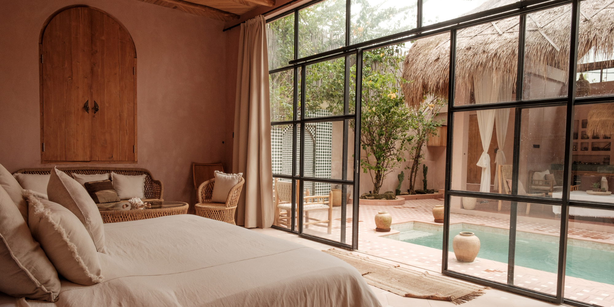 Neutral-toned bedroom with rattan chairs and a view of a private pool surrounded by tropical greenery through black-framed glass doors.