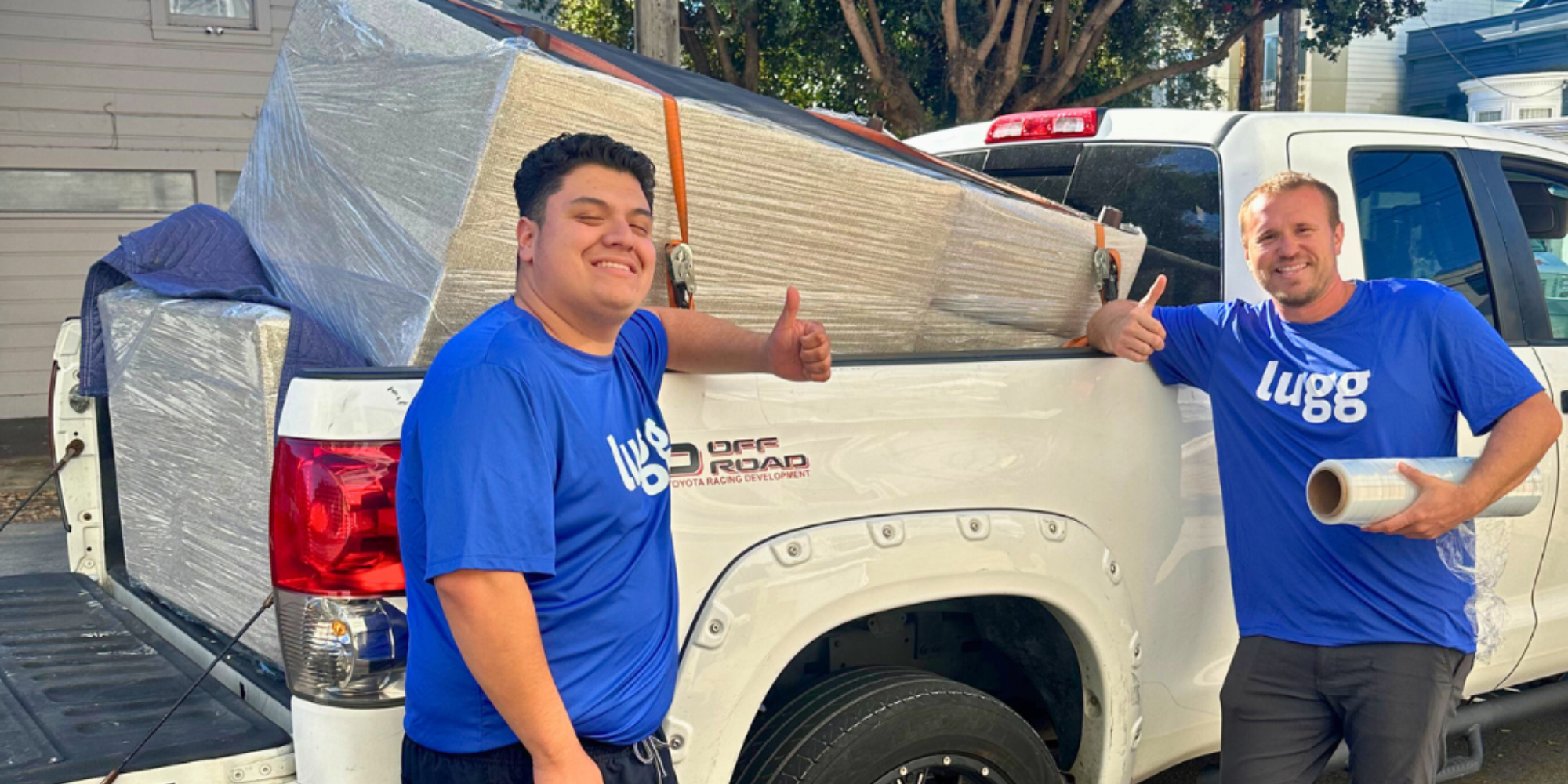 Two Lugg movers in branded blue shirts give a thumbs-up beside a white pickup truck, which holds a fully wrapped beige couch strapped in with moving pads and an orange tie-down.