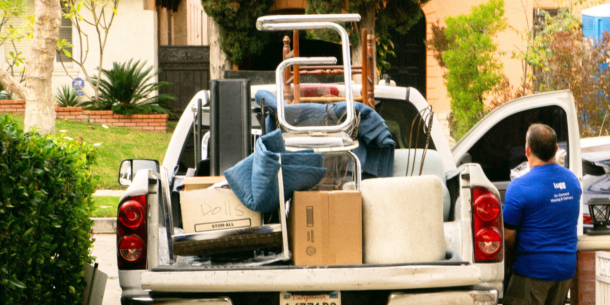 A pickup truck filled with large household items, chairs, and boxes labeled “dolls” sits parked in a driveway, with a Lugger in a blue shirt standing nearby to complete a move across town.