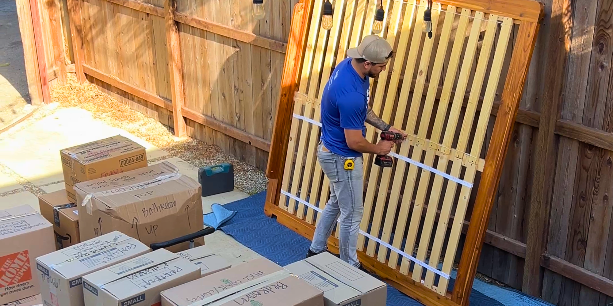 A Lugger disassembling a wooden bed frame outdoors, surrounded by labeled moving boxes, during a downsizing move.